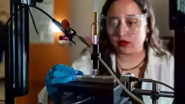 A scientist wearing safety glasses and blue gloves uses a plasma cleaner to treat a material sample.  A purple plasma beam is visible.