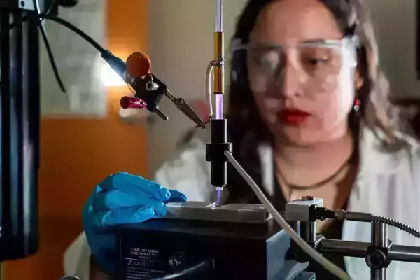 A scientist wearing safety glasses and blue gloves uses a plasma cleaner to treat a material sample.  A purple plasma beam is visible.