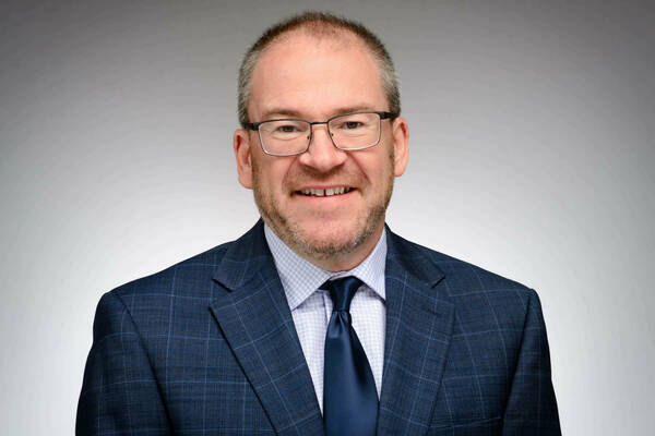 White man with glasses wearing a blue suit and tie, smiling in front of a gray background.