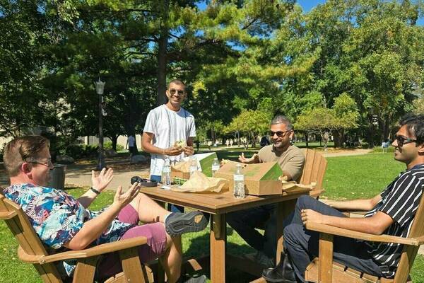 Four men sit in Adirondack chairs around a table on a sunny day on the University of Notre Dame campus, enjoying food and conversation during a Postdoc Appreciation Week event.