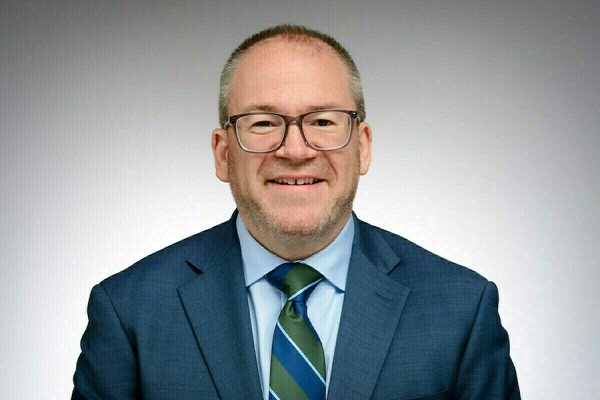 Smiling man with short gray hair, glasses, blue suit, light blue shirt, and a blue, green, and light blue striped tie.