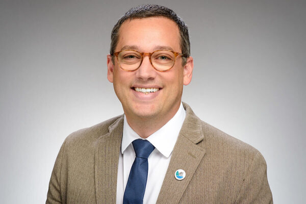 A man with graying hair, tortoise-shell glasses, blue tie, and brown herringbone blazer smiles broadly.
